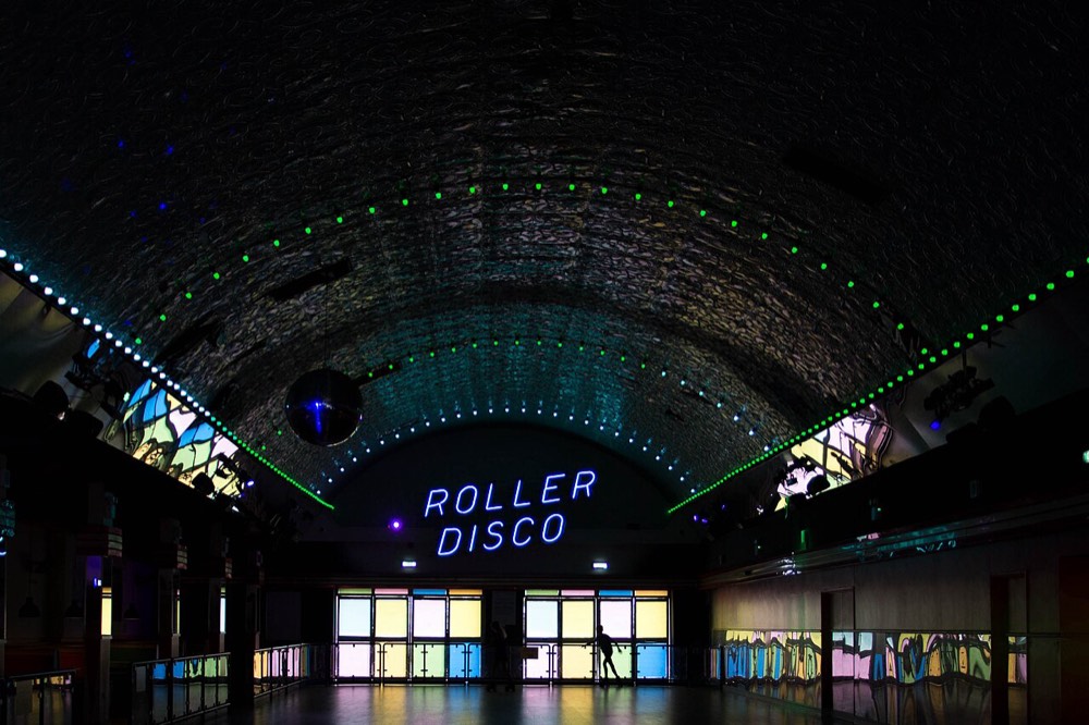 a dark roller rink hall glowing with a neon ROLLER DISCO sign and colored lights